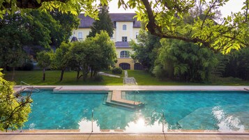 Una piscina con un camino de piedra que lleva a una fuente, rodeada de abundante vegetación y una casa grande al fondo.
