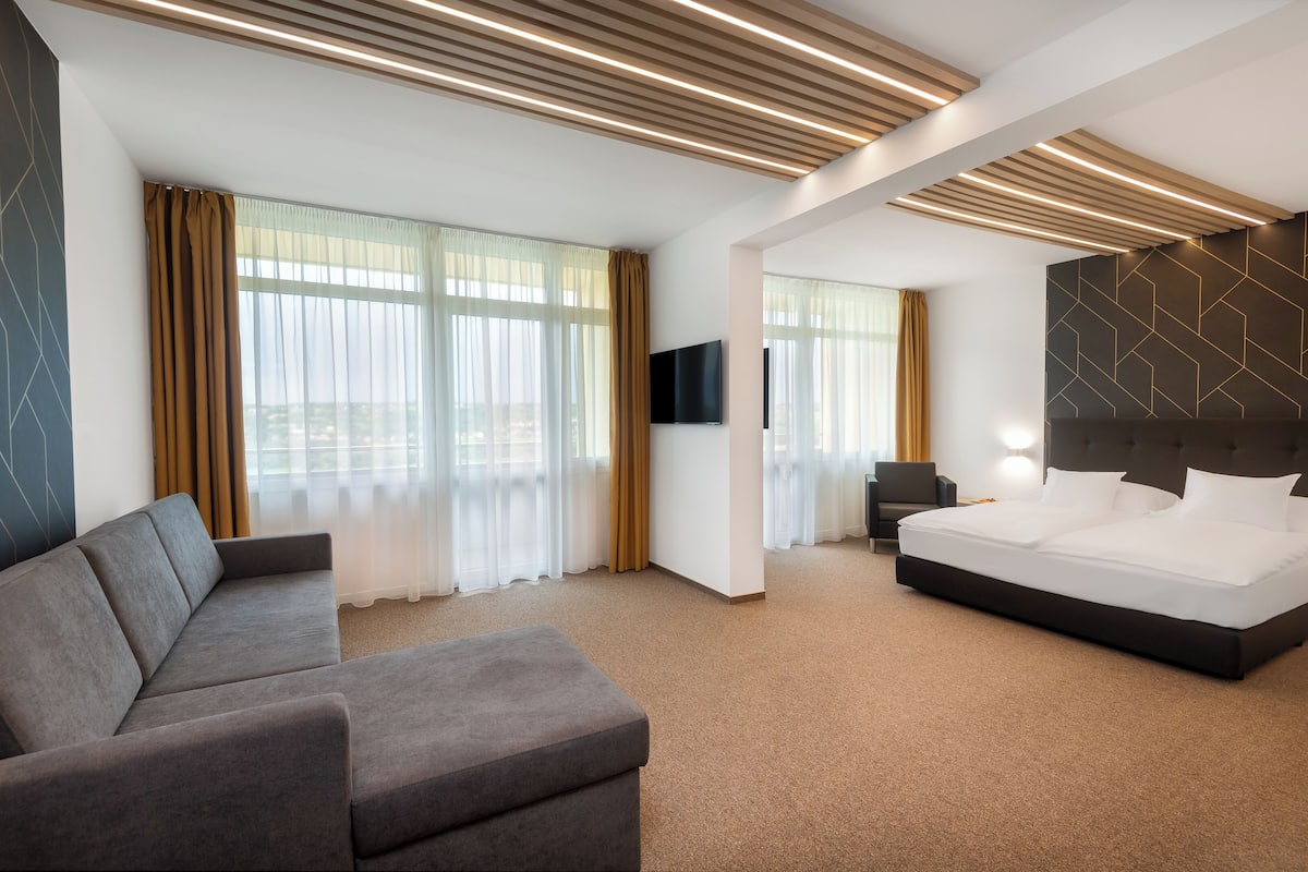 Une chambre d’hôtel moderne dotée d’un grand lit, d’un canapé, d’une télévision à écran plat et d’une grande fenêtre avec des rideaux.