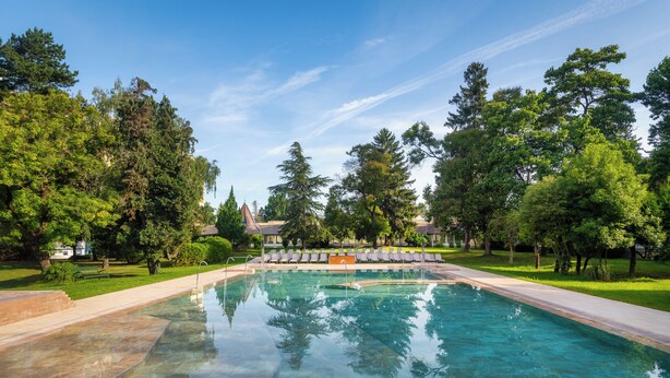 A rectangular swimming pool with clear water, surrounded by a paved deck and a grassy area with trees. - Hunguest Hotel Helios (Hévíz)