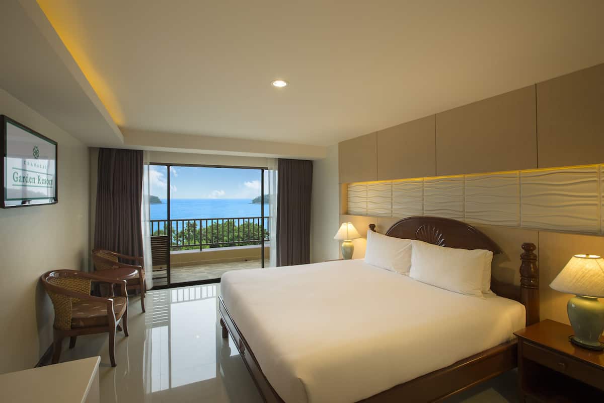 A hotel room with a large bed, two bedside lamps, a wooden headboard, a balcony with a view of the ocean, and a desk with chairs.