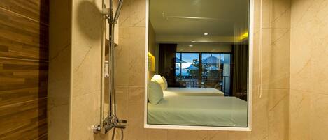 A bathroom with a bathtub filled with rose petals, a showerhead, and a mirror reflecting a bedroom with a bed and a window.
