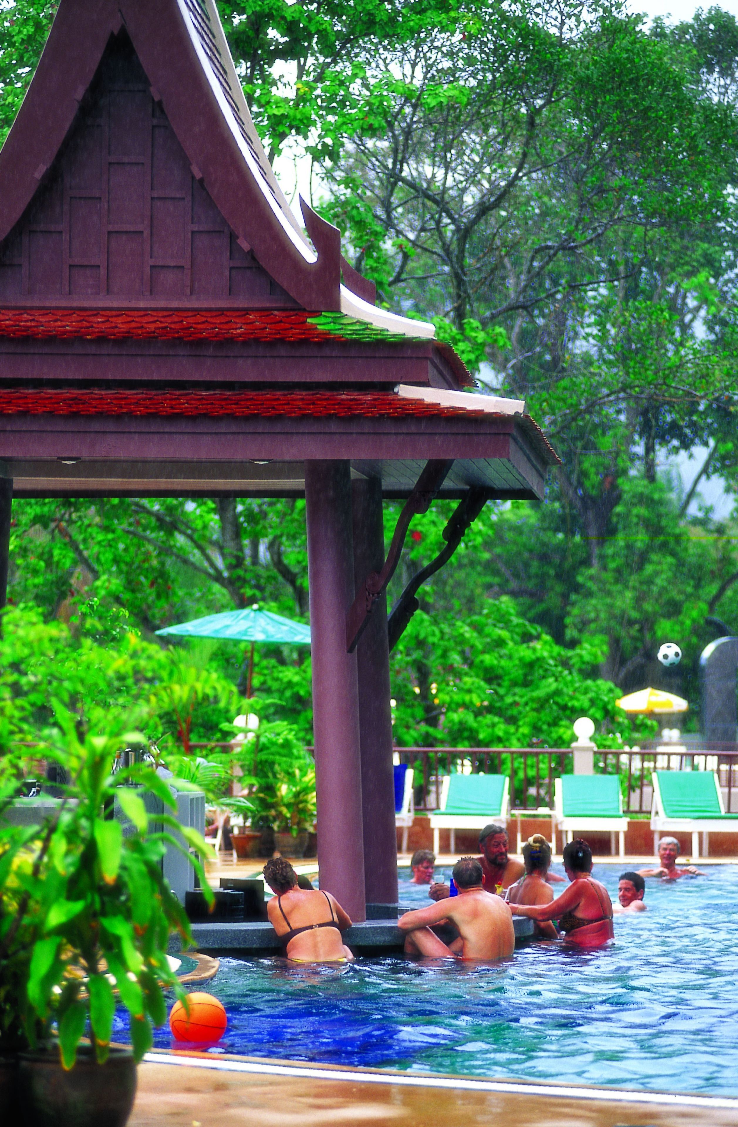 Zona de piscina con una estructura de pabellón tradicional, sillas de descanso y abundante vegetación de fondo.