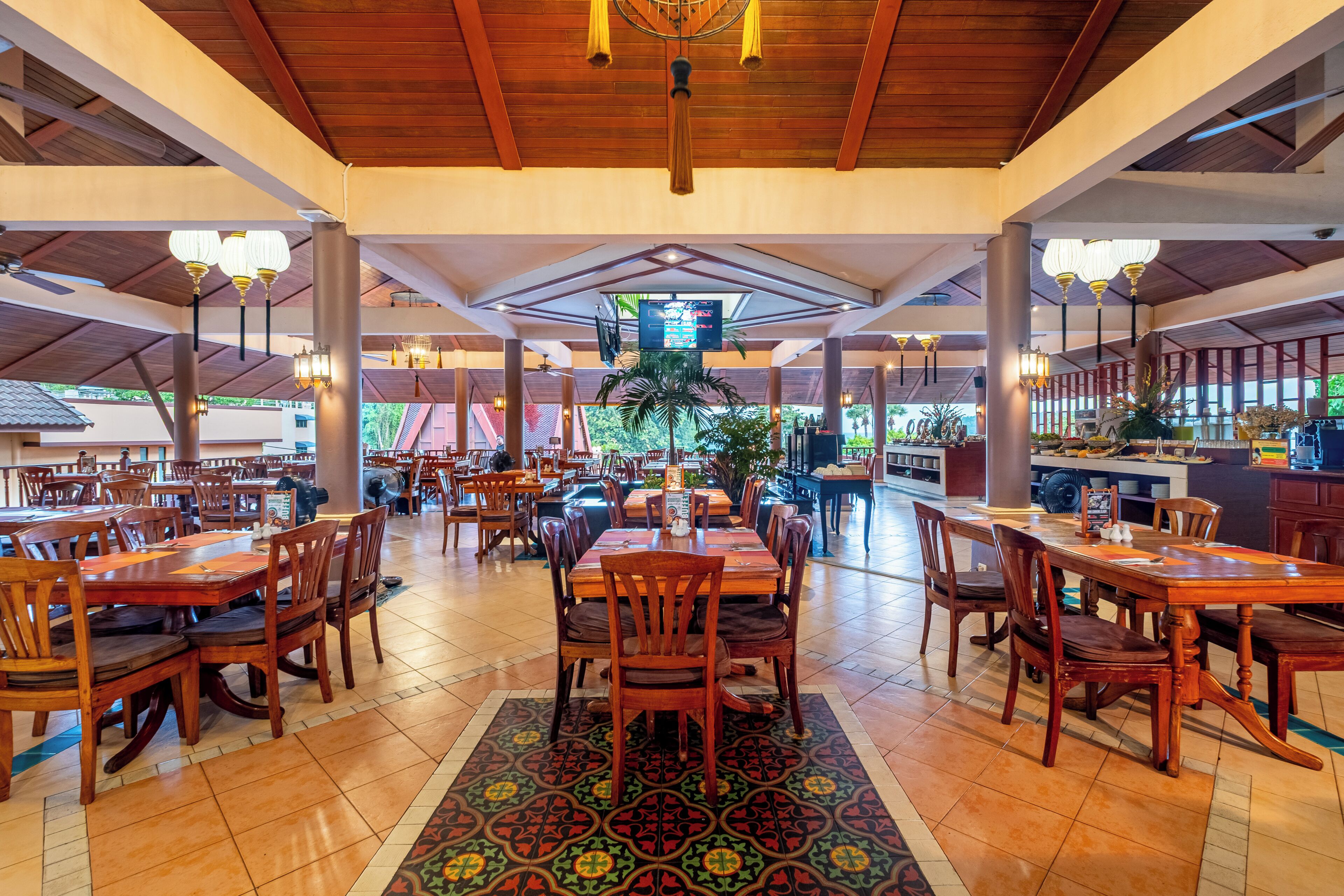 A spacious restaurant with wooden tables and chairs, a patterned rug, and a ceiling with hanging lights.