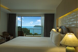 A hotel room with a large bed, a nightstand, a lamp, and a view of the sea through a balcony.
