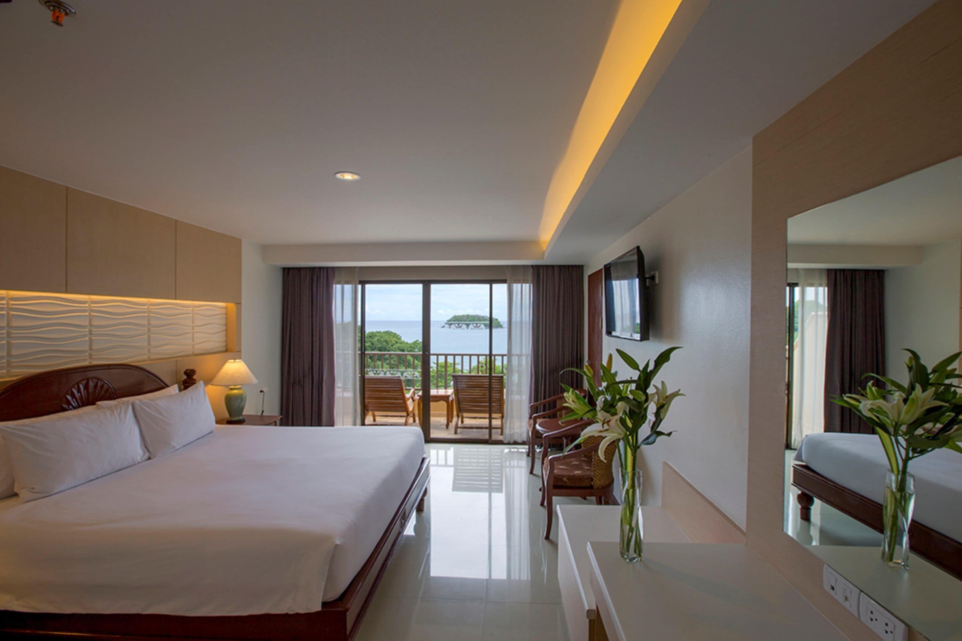 A hotel room with a large bed, a desk with a chair, a TV mounted on the wall, and a balcony with a view of the sea.