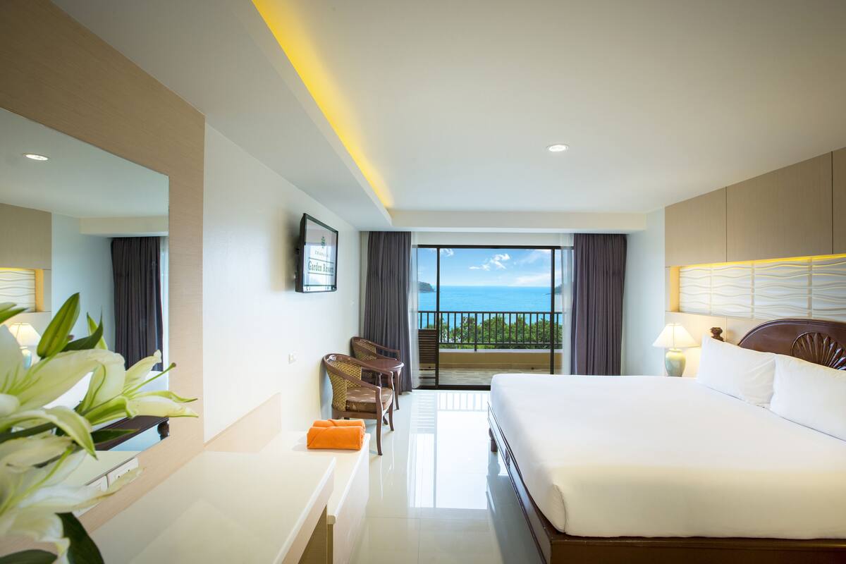 A hotel room with a large bed, a desk with a chair, a small table with a chair, a balcony with a view of the ocean, and a framed picture on the wall.