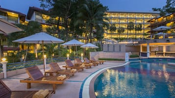 A poolside area with lounge chairs and umbrellas, a swimming pool, and a hotel building in the background.