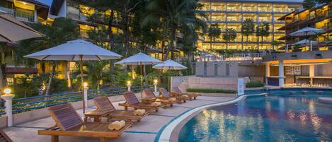 A poolside area with lounge chairs and umbrellas, a swimming pool, and a hotel building in the background.