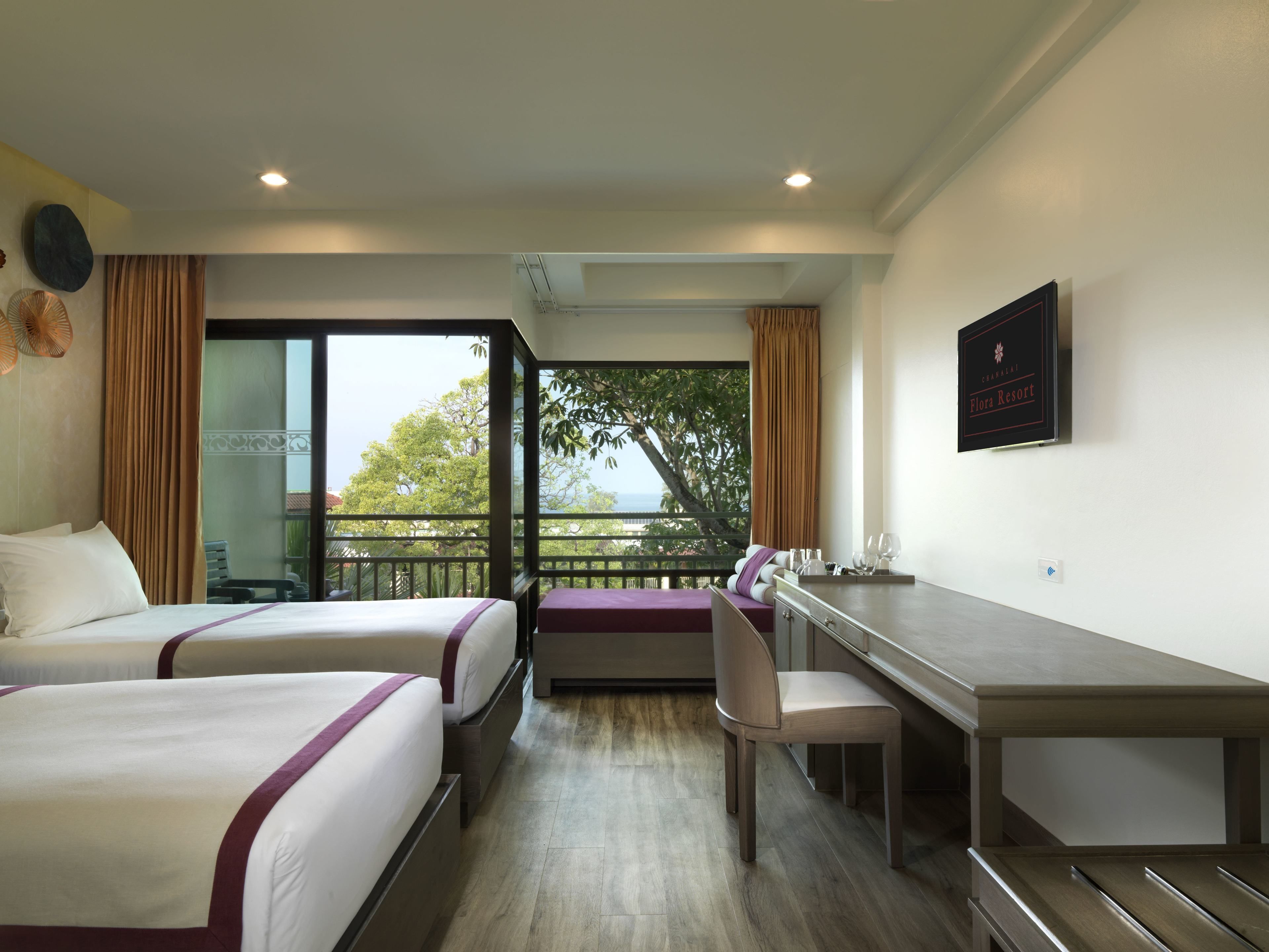 A hotel room with two beds, a desk, and a chair. There is a balcony with a view of trees and a building.
