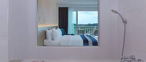 Baño de hotel con una bañera llena de pétalos de rosa, ducha y un espejo grande que refleja un dormitorio con cama y vistas a un balcón.
