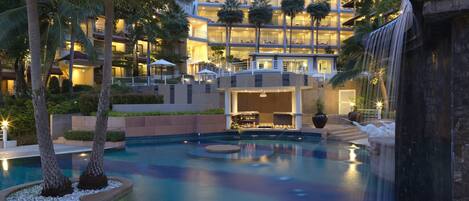 A hotel with a swimming pool, palm trees, and a waterfall.