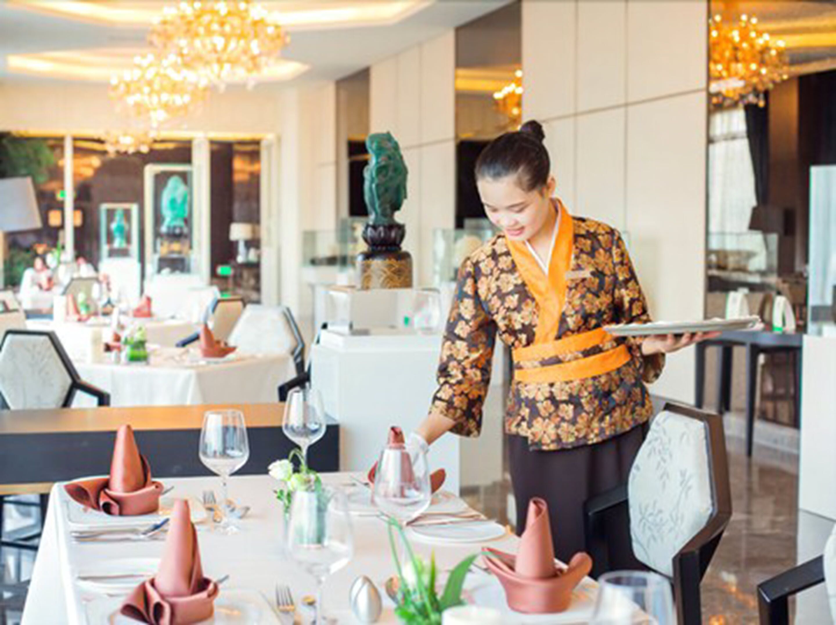 A well-lit dining area with tables set for a meal, a waiter in traditional attire, and elegant decor including a chandelier and a statue.