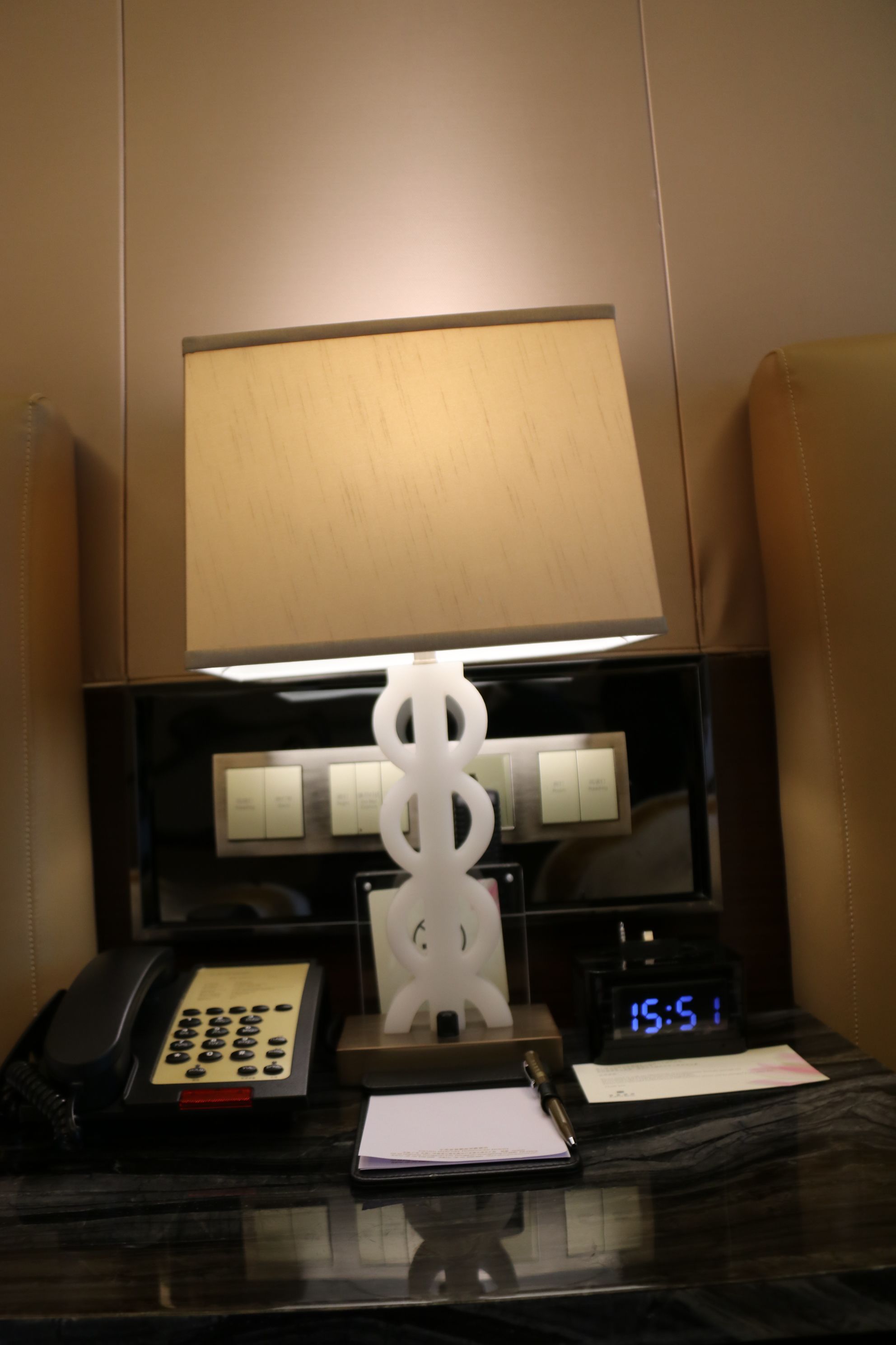 A hotel room with a desk featuring a telephone, a notepad, a pen, a clock, and a lamp.