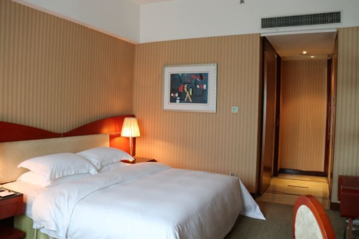 A hotel room with a bed, a nightstand, a lamp, a framed picture on the wall, and a door leading to another room.