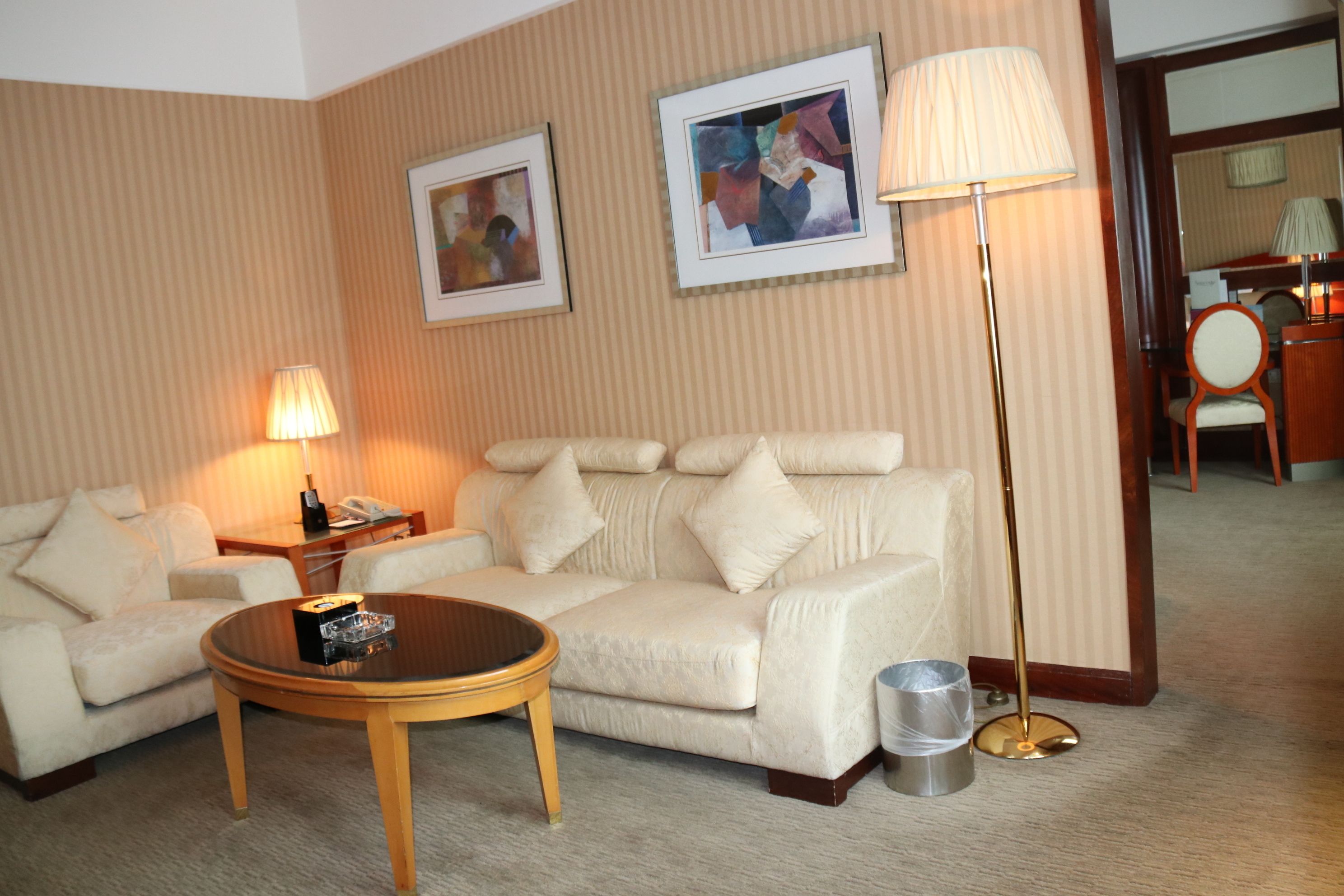 A hotel room with a beige sofa, a round coffee table, a lamp, a trash can, and two framed pictures on the wall.
