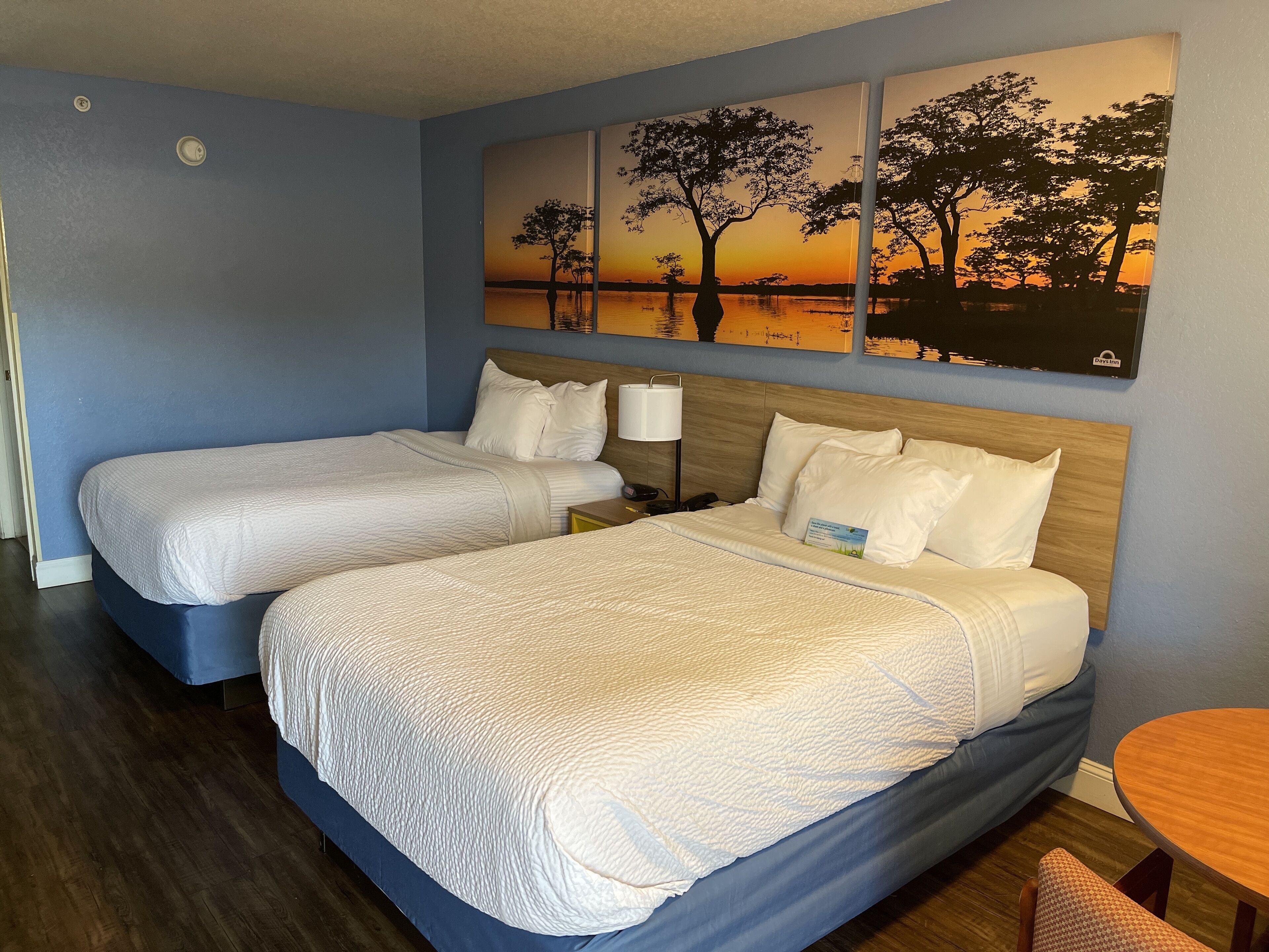 A hotel room with two beds, a wooden headboard, a painting of trees at sunset, a small round table, and a chair.