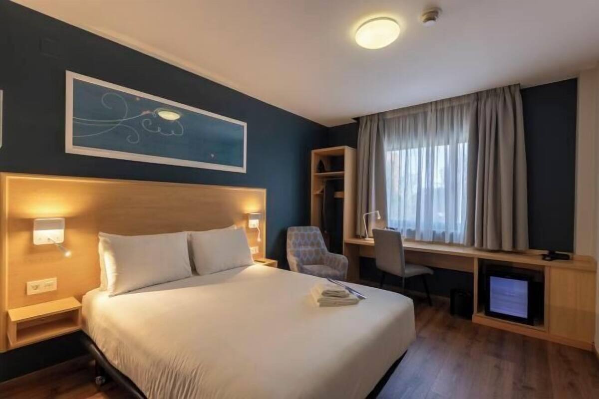 Ein Hotelzimmer mit einem großen Bett, einem Schreibtisch, einem Sessel, einem Fernseher und einem Fenster mit Vorhängen.