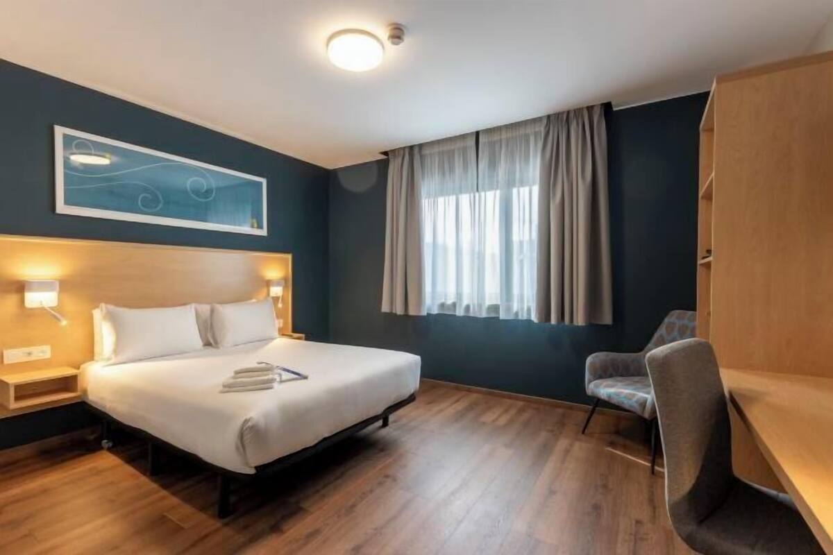 Ein Hotelzimmer mit einem Bett, Nachttischen, einem Sessel, einem Fenster mit Vorhängen und einem Wandbild.