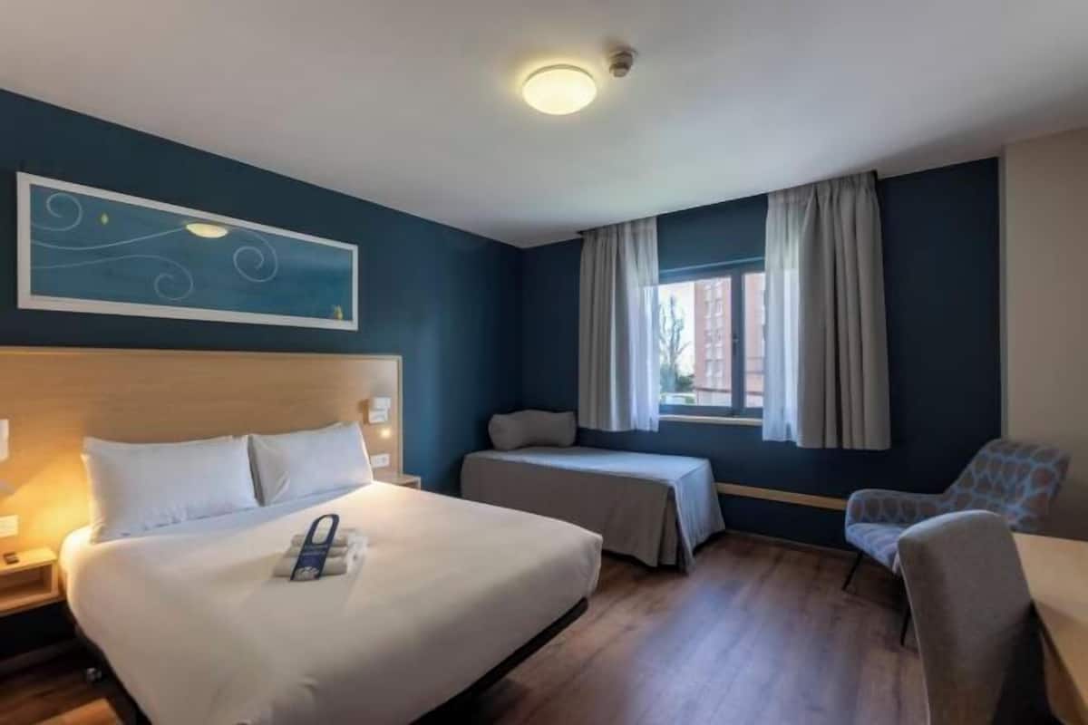 Ein Hotelzimmer mit einem großen Bett, zwei Einzelbetten, einem Schreibtisch, einem Stuhl und einem fenster mit Vorhängen.