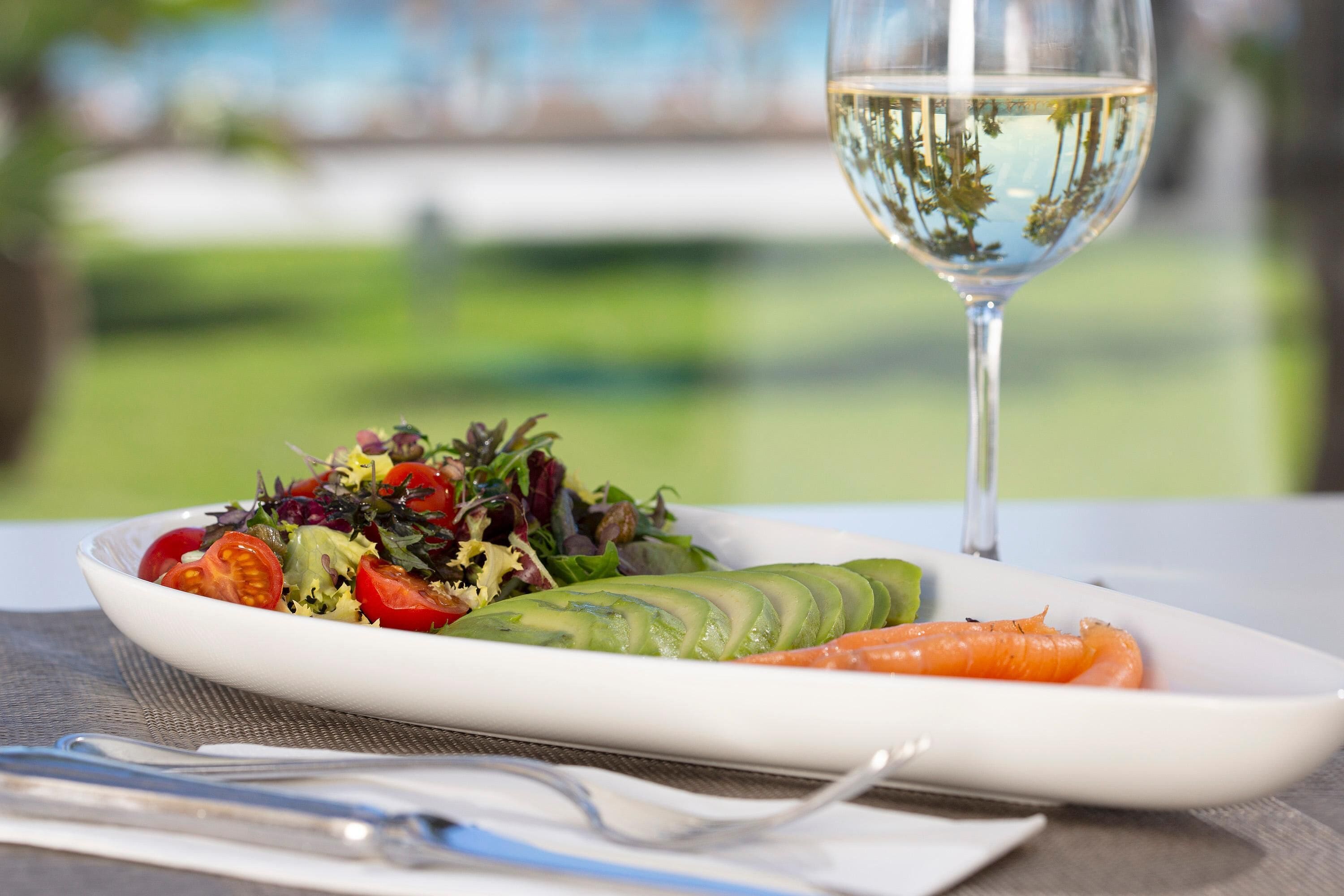 Un plato blanco con ensalada, una rebanada de aguacate y una zanahoria, acompañado de una copa de vino blanco sobre una mesa.