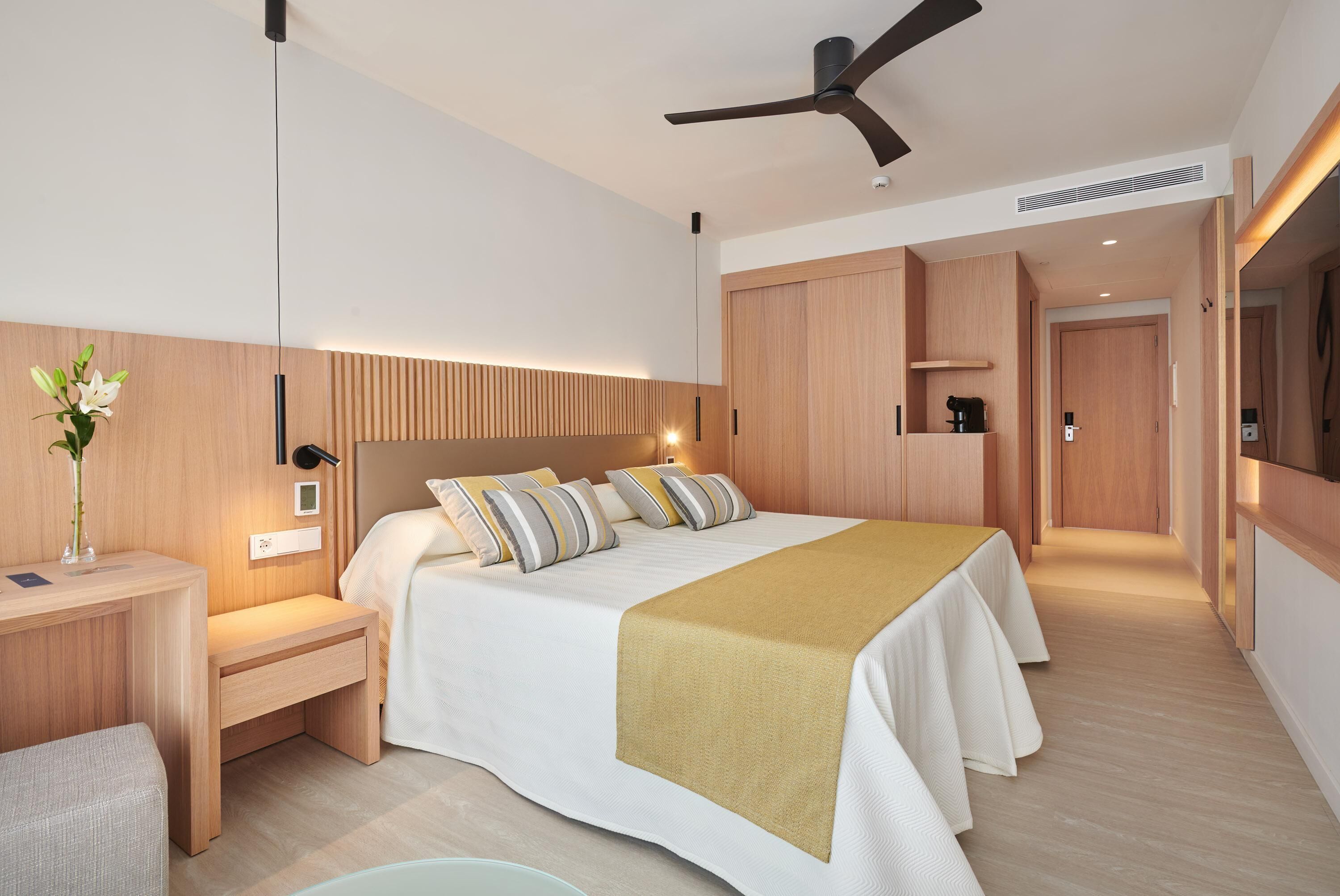 Una habitación de hotel moderna con una cama grande, mesitas de noche, un escritorio, un ventilador de techo y un armario de madera.
