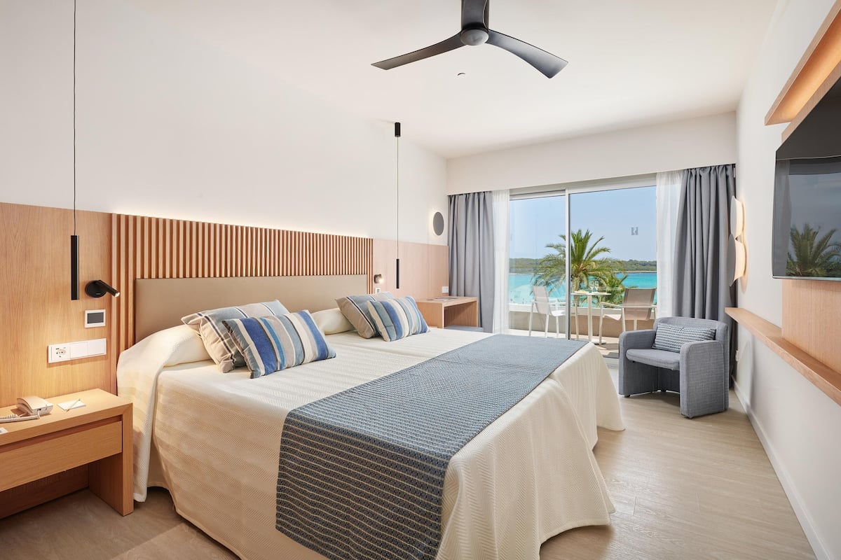 Ein modernes Hotelzimmer mit einem großen Bett, einem Schreibtisch, einem Sessel und einem Balkon mit Meerblick.