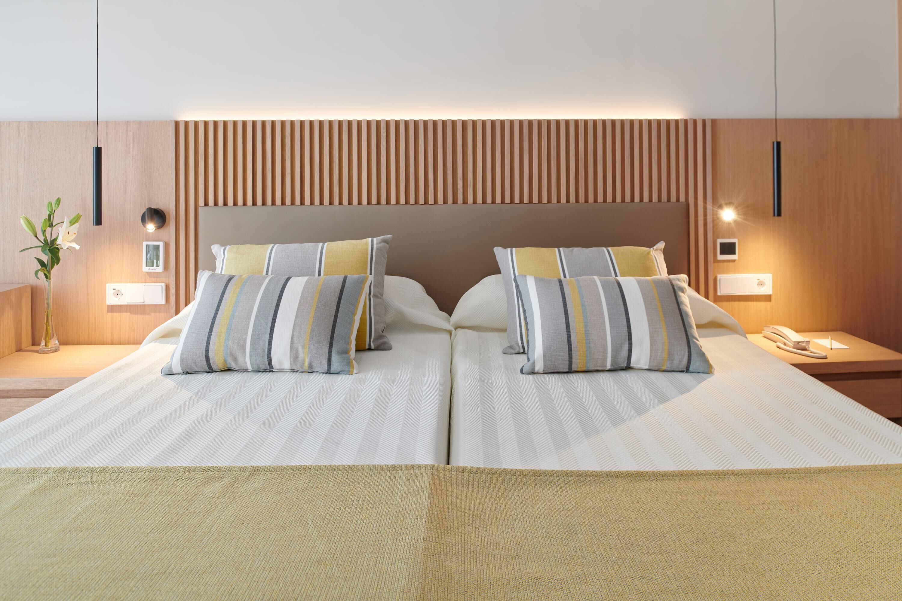 Una cama doble con ropa de cama a rayas, cabecera de madera, mesitas de noche con lámparas y un florero con flores.