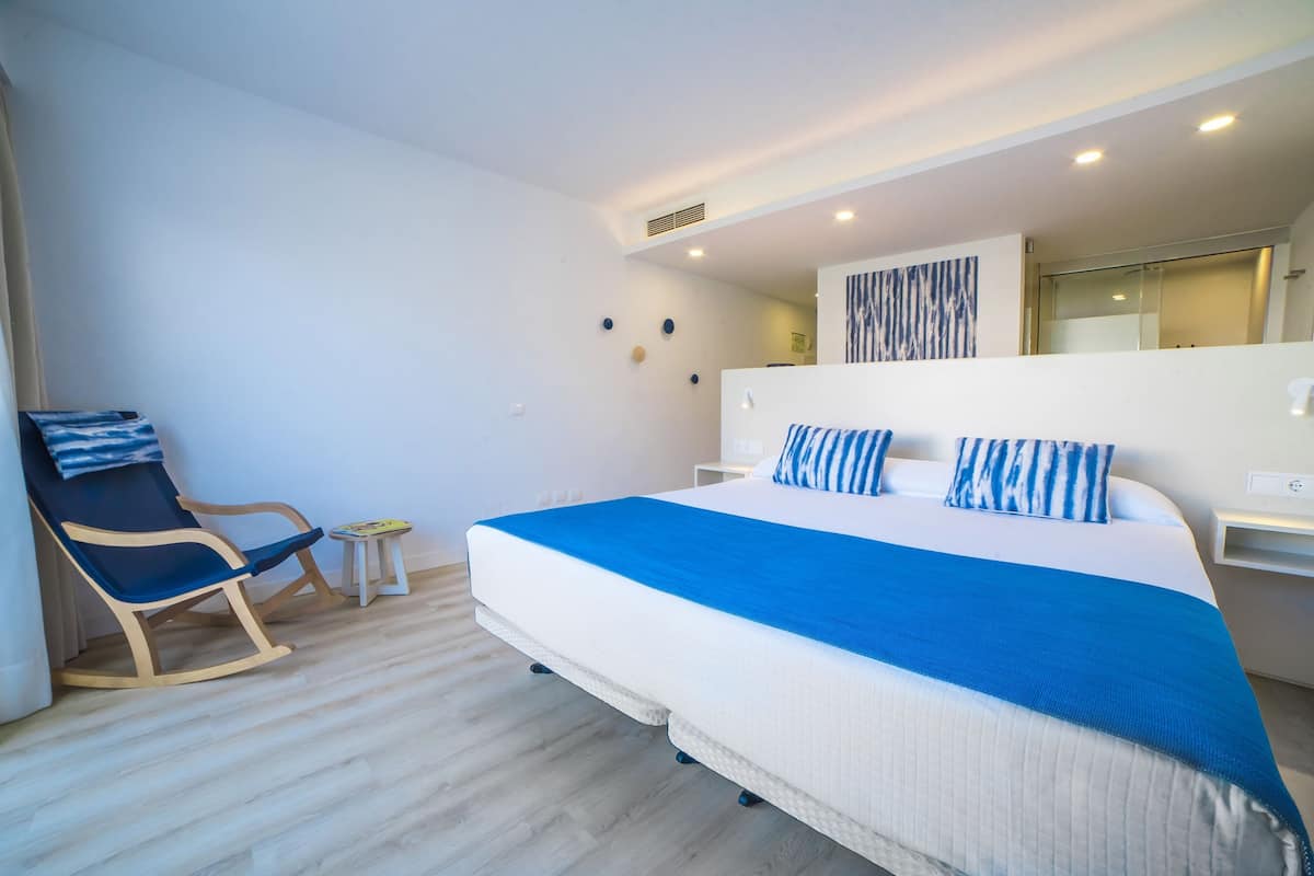 A modern hotel room with a large bed, a rocking chair, a small table, and a wall-mounted TV.