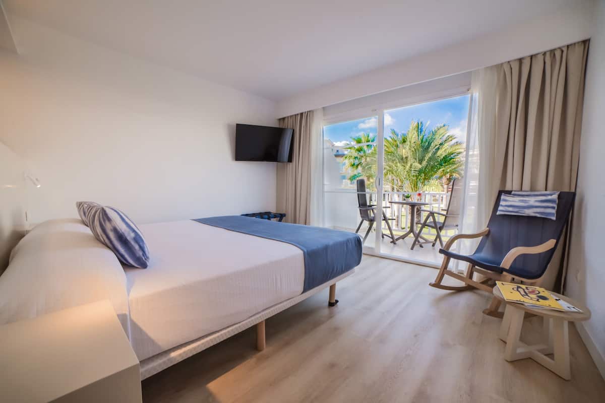A hotel room with a bed, a TV, a balcony with a table and chairs, and a view of palm trees.