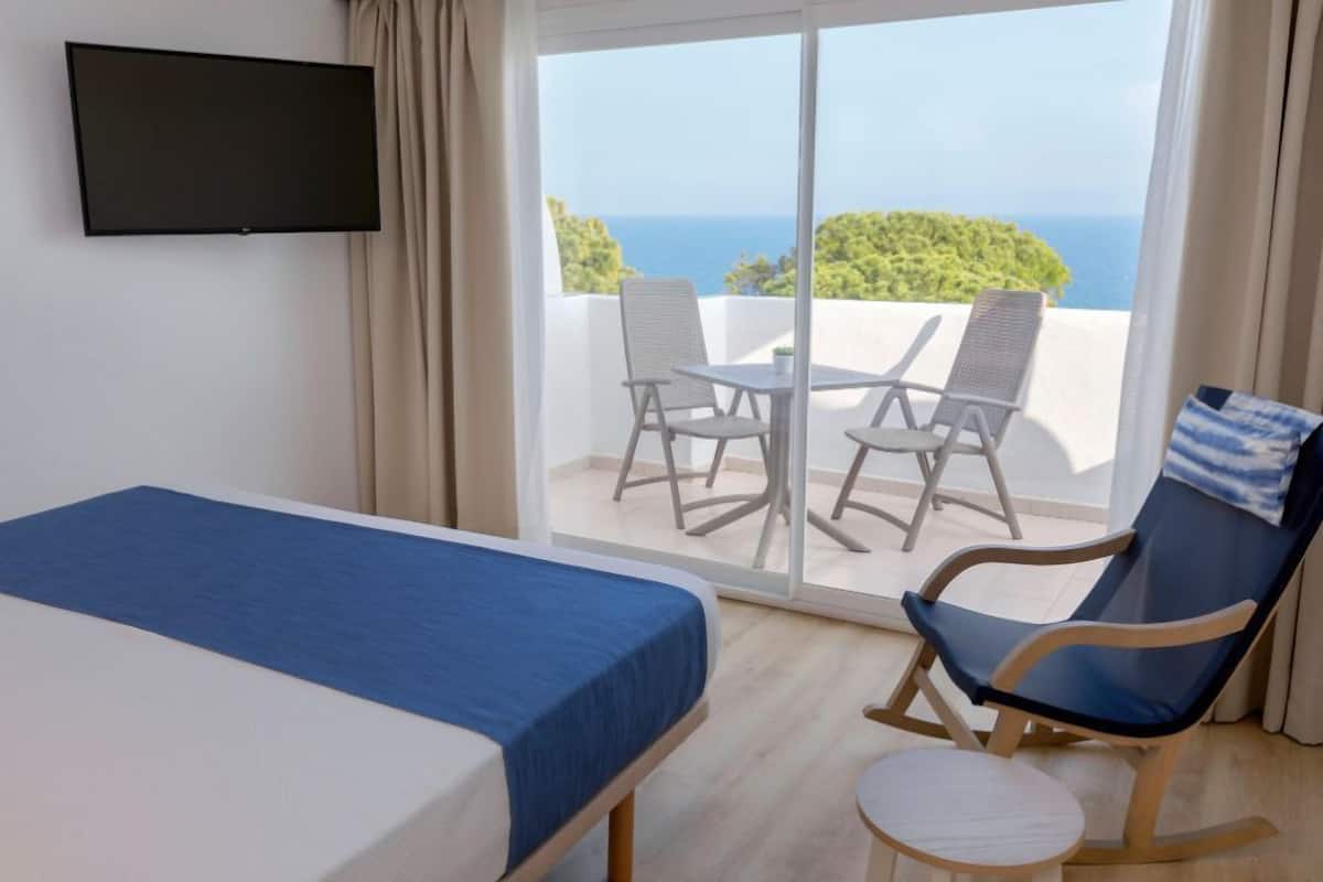 A hotel room with a bed, a TV, a balcony with a table and chairs, and a view of the sea.