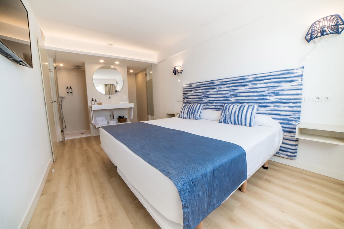A modern bedroom with a large bed, a bedside table, a mirror, and a shower area.