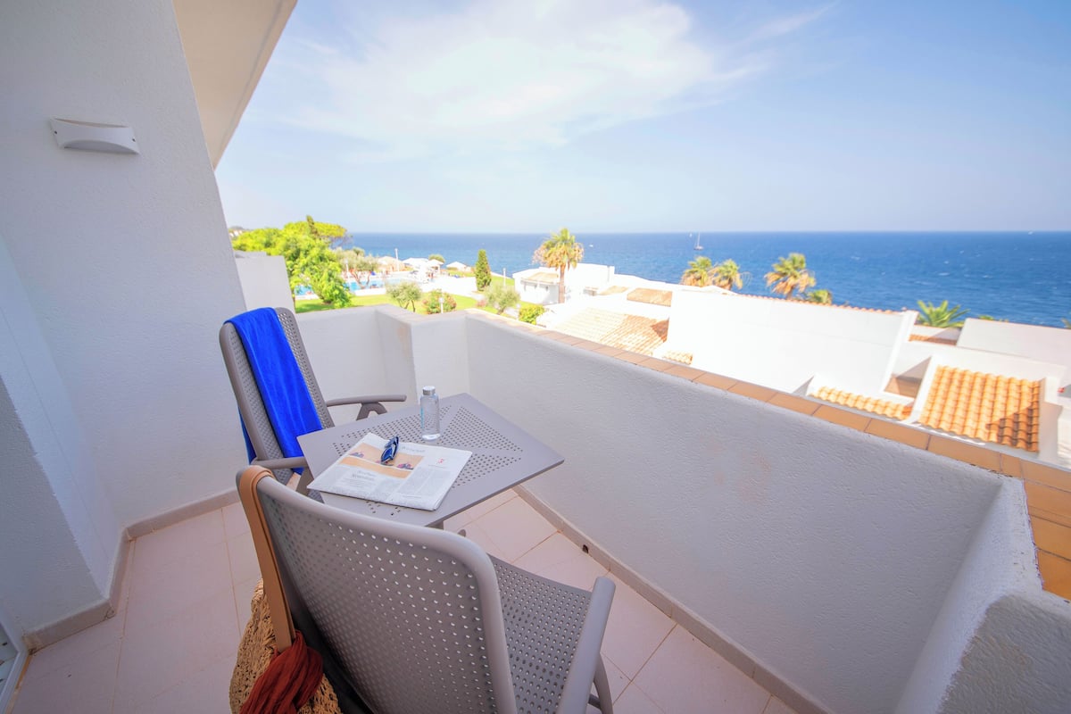 A balcony with a table and chairs, overlooking a coastal view with buildings and palm trees.