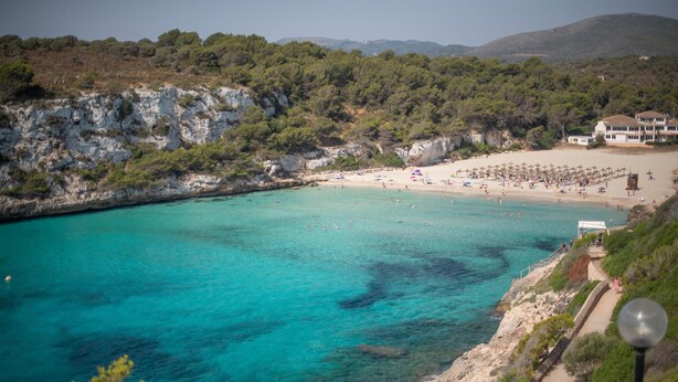 A coastal beach with clear turquoise waters, a rocky shoreline, and a path leading to a beach area with umbrellas and people. - Blau Punta Reina (Manacor)