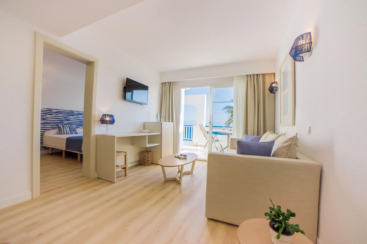 A modern living room with a sofa, a coffee table, and a TV mounted on the wall. There is a balcony with a view of the sea.