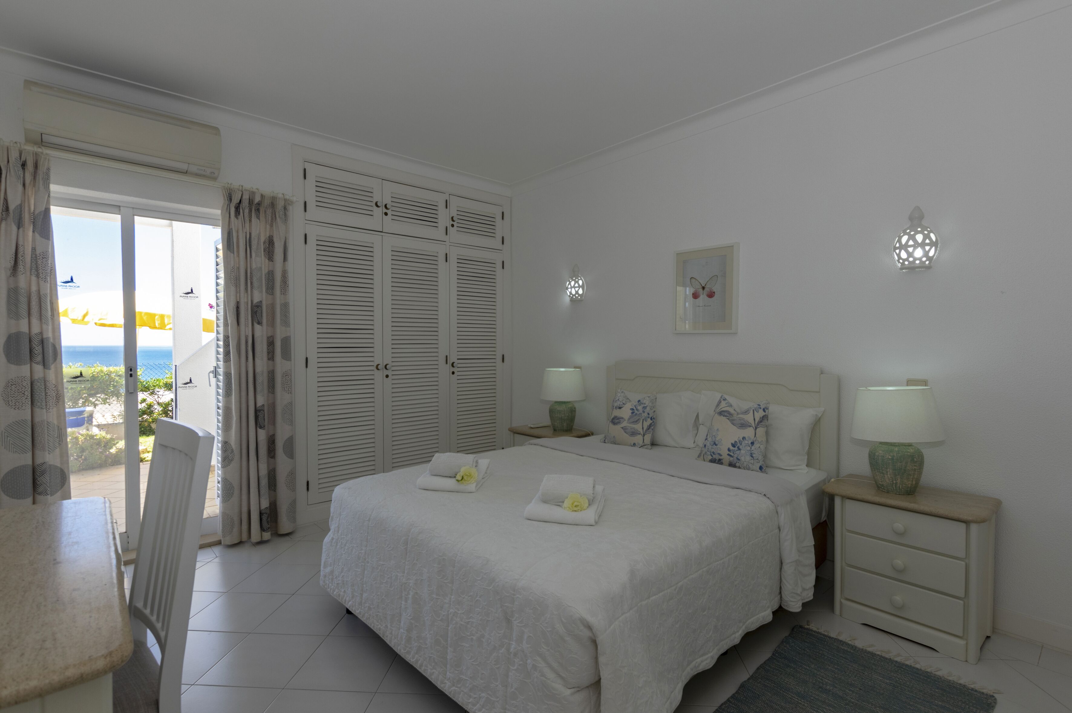 A bedroom with a bed, nightstands, a chair, a desk, and a view of the outside.