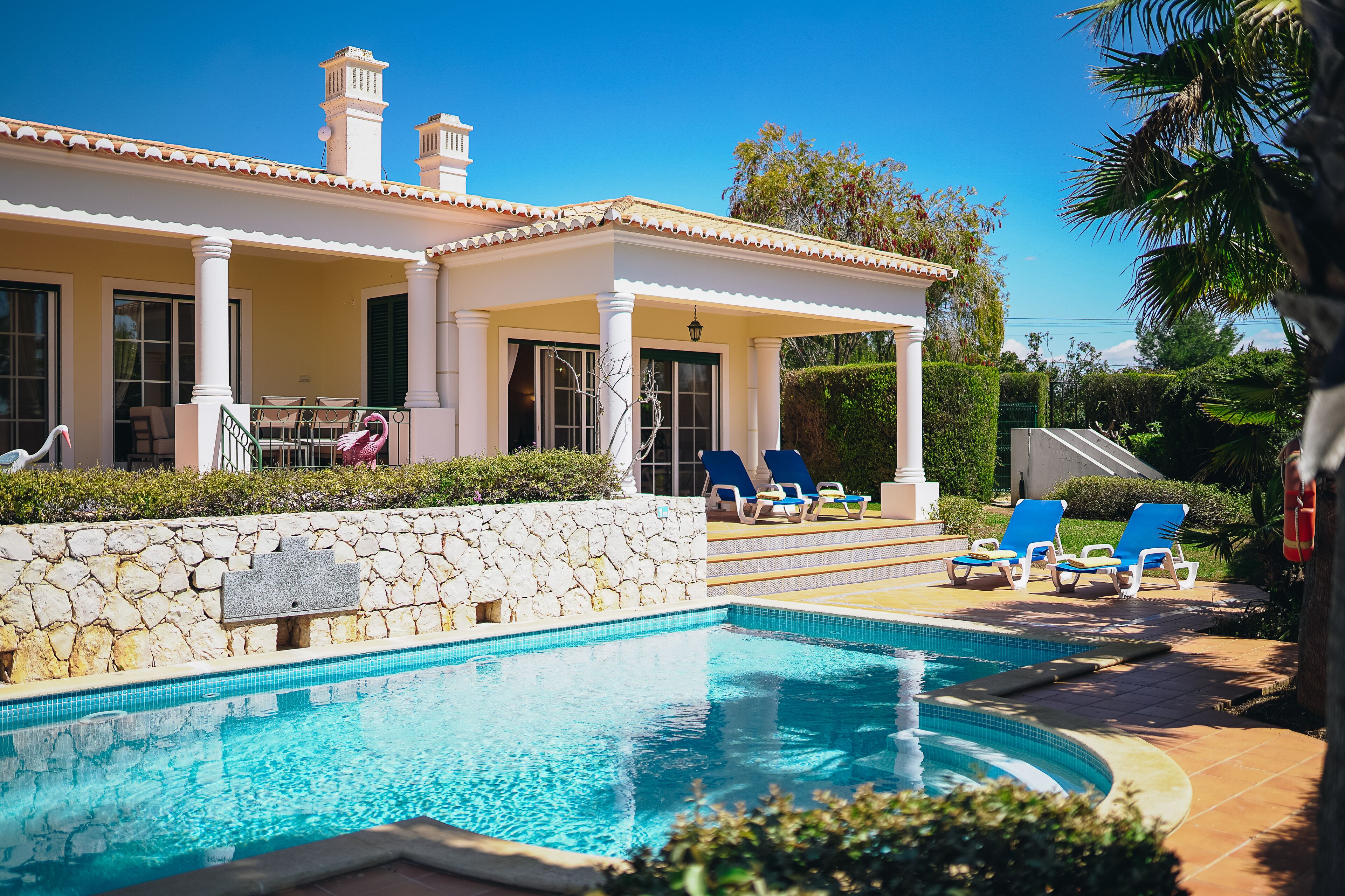 A pool area with a stone wall, a covered seating area with chairs, and a clear blue pool.