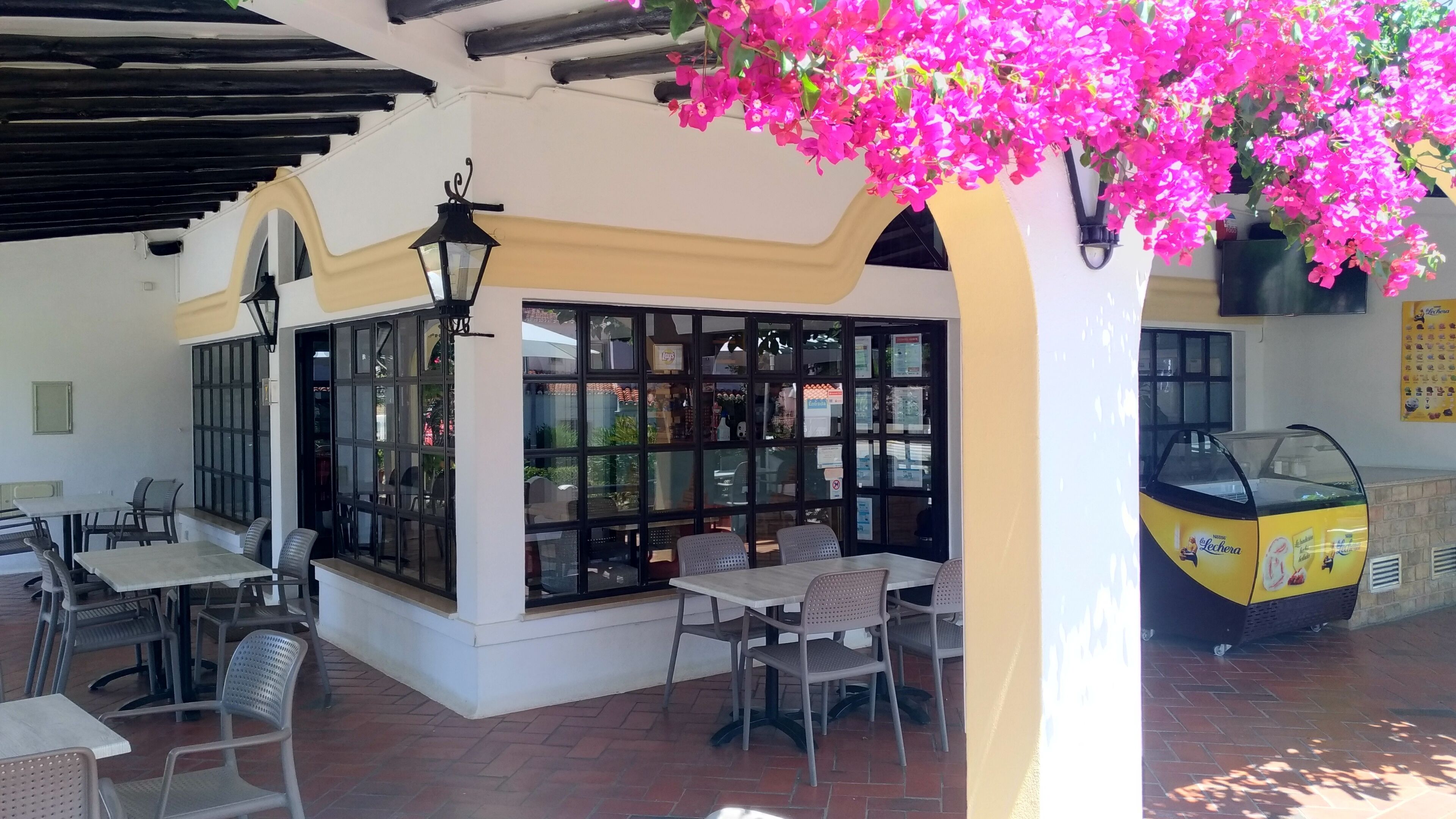 Un café al aire libre con mesas y sillas, una máquina expendedora amarilla y un cerramiento de vidrio con vistas a la calle.