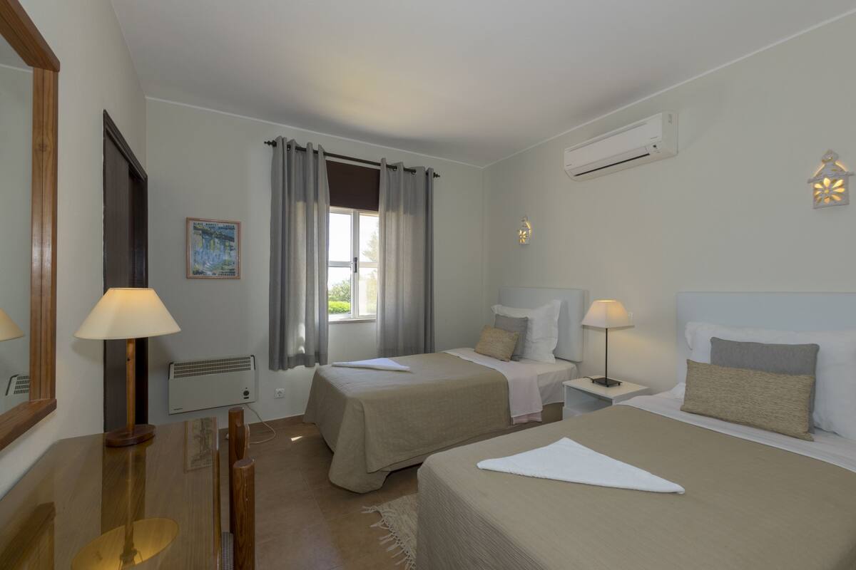 A hotel room with two beds, a window with curtains, a wall-mounted air conditioner, a nightstand with a lamp, and a framed picture on the wall.