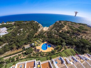 An aerial view of a coastal property with a swimming pool, lush greenery, and a clear view of the sea.