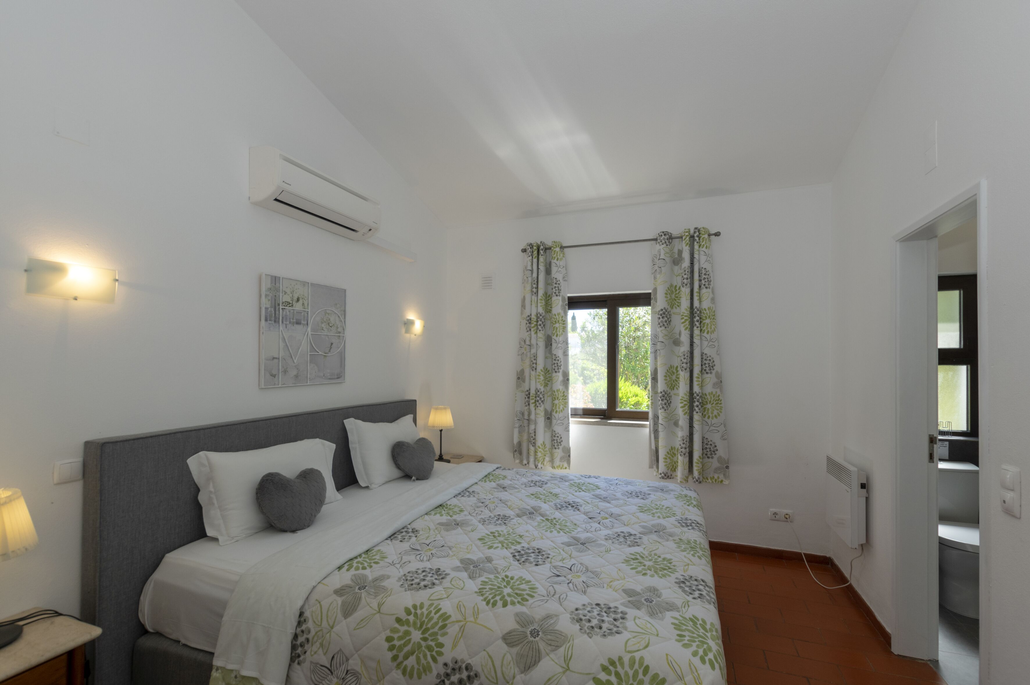 Een slaapkamer met een bed, nachtlampjes, een airconditioner, een raam met gordijnen en een kunstwerk aan de muur.