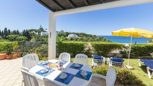 A covered outdoor dining area with white chairs and a table set for four, overlooking a scenic view of the sea and hills. - Rocha Brava Village Resort (Lagoa)