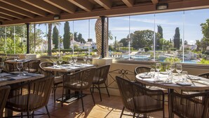 An outdoor dining area with tables and chairs, large windows, and a view of a pool and greenery. - Rocha Brava Village Resort (Lagoa)
