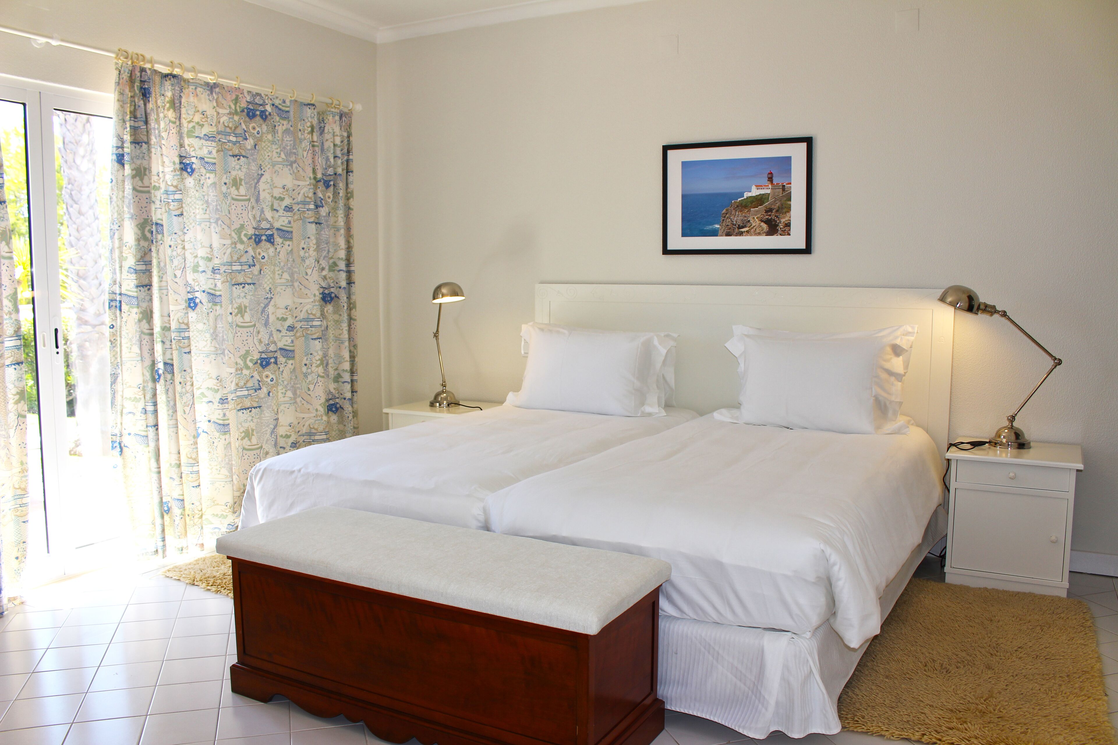 A double bed with white linens, a wooden bench, two bedside tables with lamps, a framed picture on the wall, and a large window with curtains.