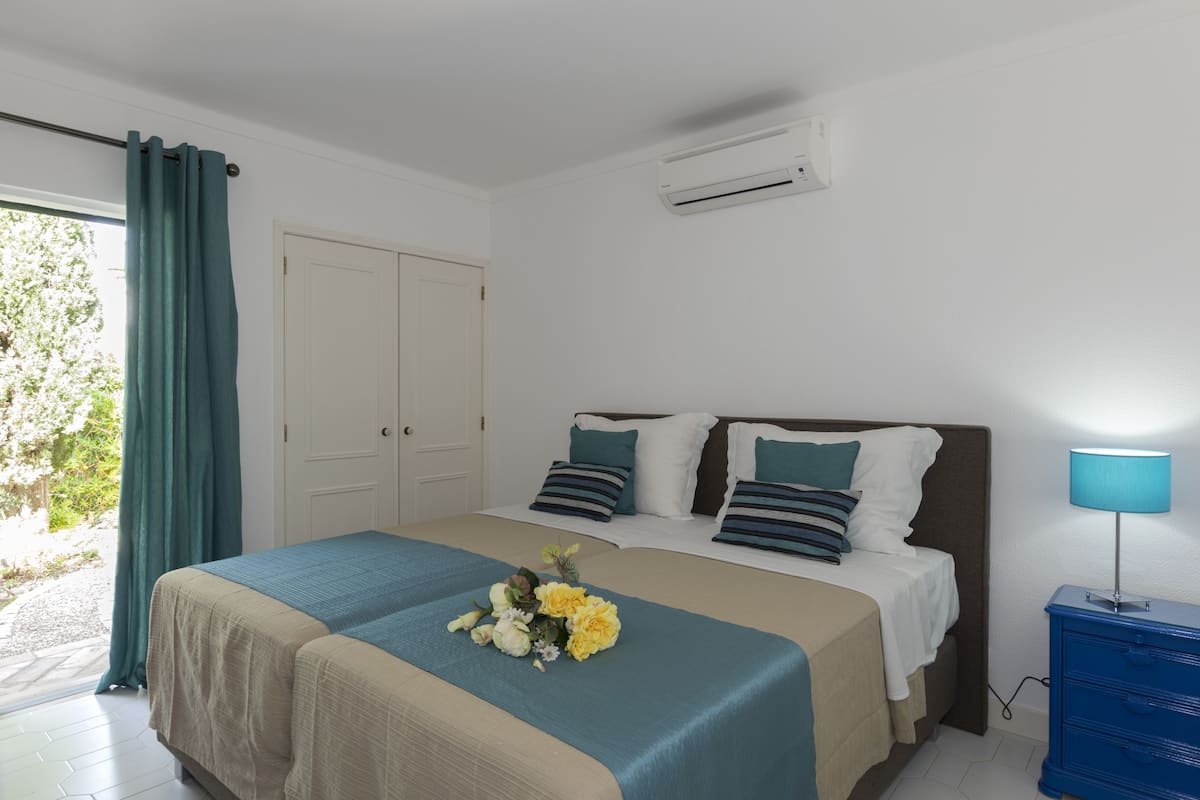Une chambre à coucher comprenant un lit, une table de chevet, une lampe, une fenêtre avec des rideaux et un appareil de climatisation.