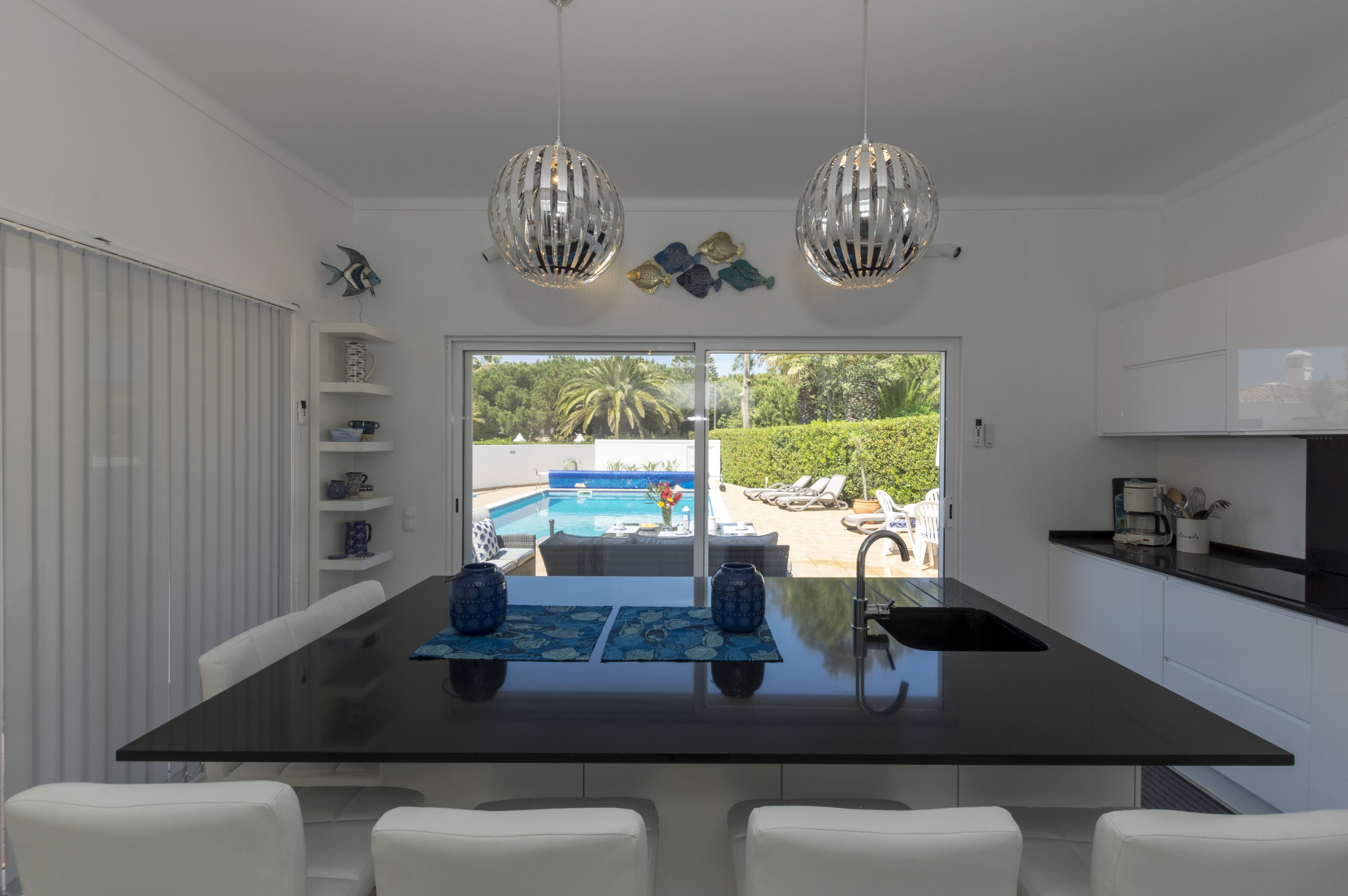 A modern kitchen with a central island, white cabinetry, and pendant lighting. A large window offers a view of a pool and greenery.