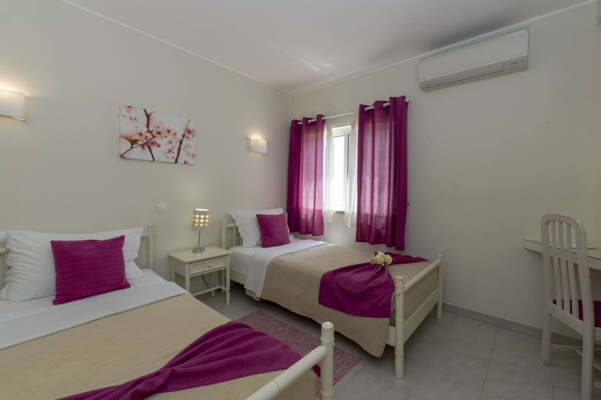 Une chambre d’hôtel avec deux lits, un bureau, une chaise, une fenêtre avec des rideaux violets, un climatiseur et un luminaire fixé au mur.