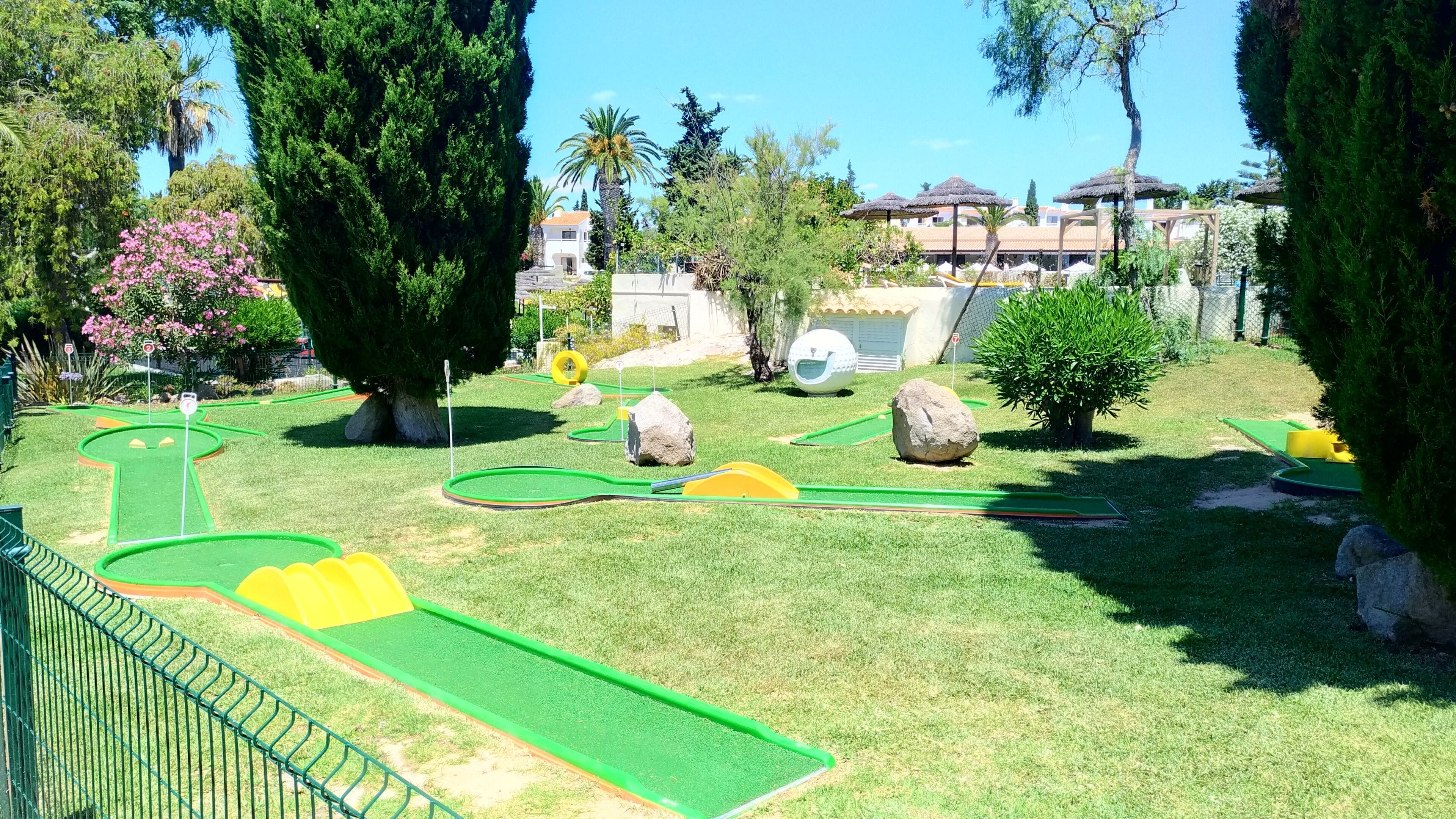 A miniature golf course with green and yellow holes, surrounded by trees and a fence.