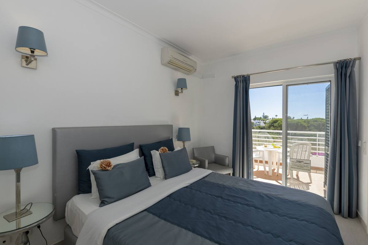 A hotel room with a bed, bedside tables, a lamp, an air conditioner, and a balcony with outdoor furniture.