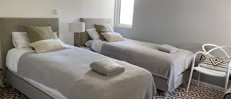 Habitación de hotel con dos camas, piso estampado, una ventana con persianas y una silla blanca.