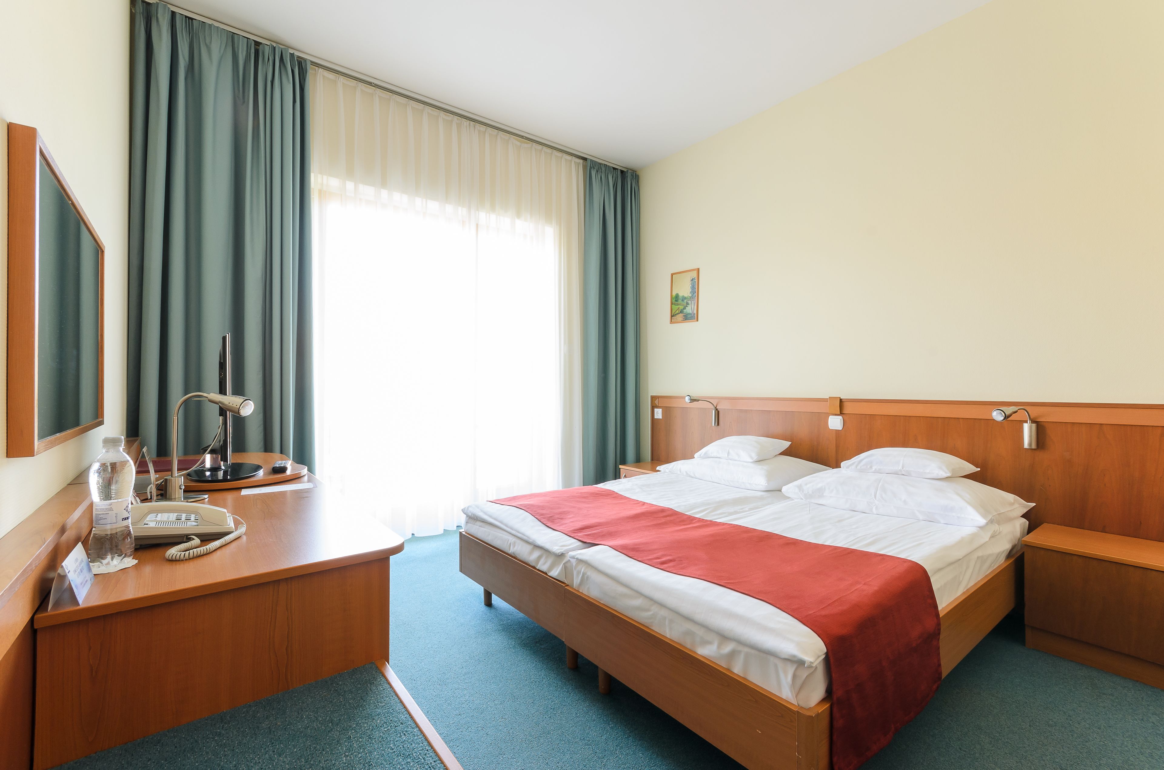 A hotel room with a wooden bed, a desk with a lamp, a telephone, and a window with curtains.